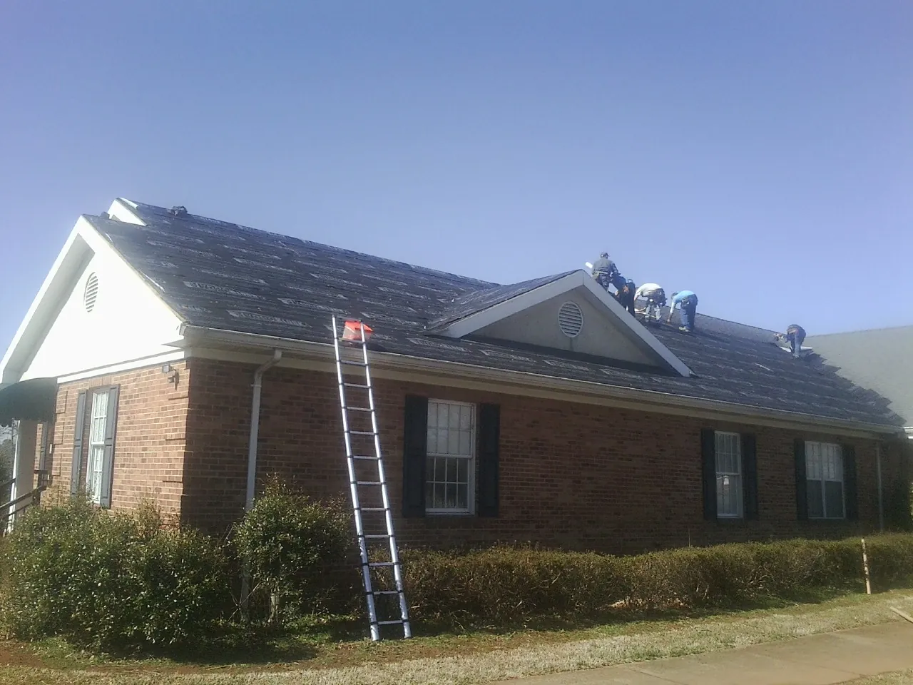 Crew tearing shingles off a brick ranch roof with a ladder set — Emergency Roof Repair work by our crew in Misenheimer