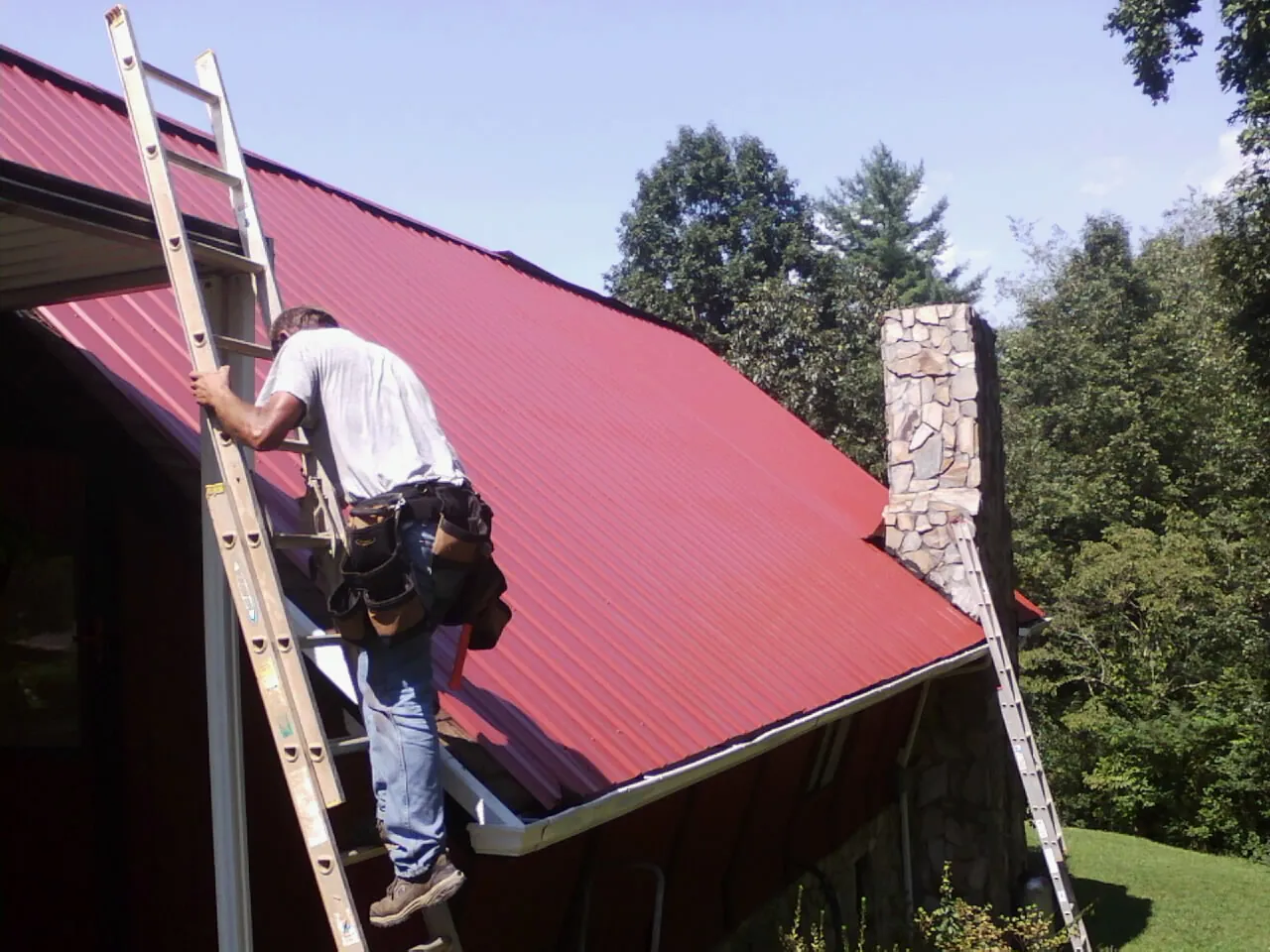 Roofer on a ladder beside a red metal roof and stone chimney — Soffit & Fascia Repair by Roofing Brothers in Misenheimer